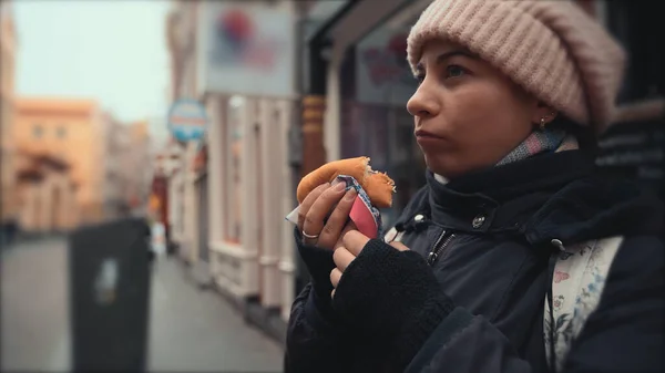 Pelancong wanita muda yang kelaparan makan makanan cepat saji tanpa daging di jalanan dengan latar belakang kota Eropa yang kabur — Stok Foto