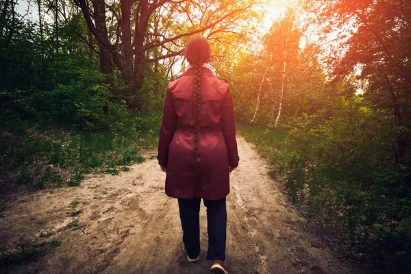Ung kvinna i röd jacka eller mantel och flätade hår promenader i vårskogen på gångväg, bakifrån — Stockfoto