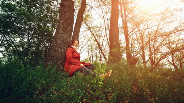 Ung söt kvinna i röd rock med vila i vårskogen och tittar på kameran — Stockfoto