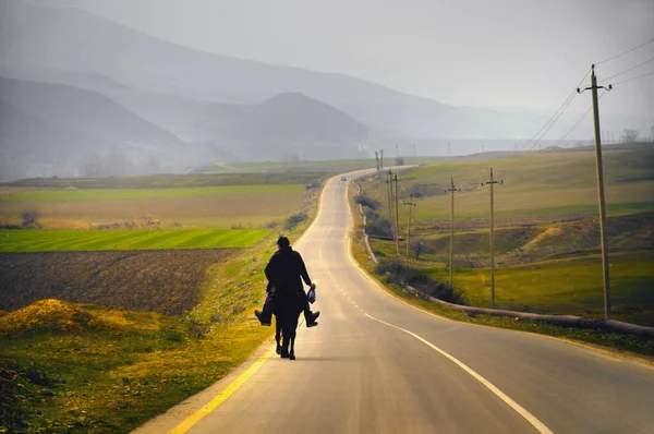 Paisagem de um homem montando um cavalo ao longo da estrada de asfalto entre campos verdejantes em um dia de primavera claro e com as montanhas na distância — Fotografia de Stock
