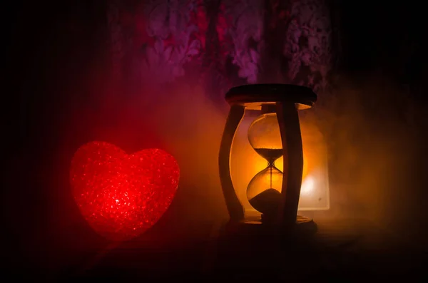 バーニング暗い背景と砂時計シルエット時計、時間と愛の概念、セレクティブ フォーカスや愛グリーティング カード中心の赤い緑の色とりどりのハートの甘いバレンタインの組成 — ストック写真