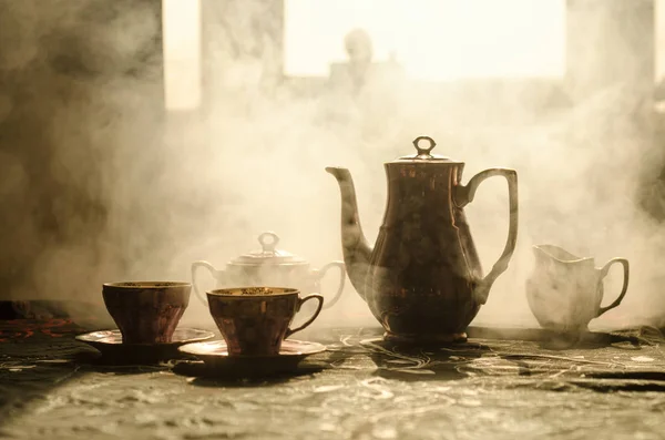 Comida fondo té y café tema. Tetera de cerámica antigua o cafetera con jarra de tazas y taza de azúcar sobre fondo oscuro con luces y humo. Siluetas de tetera y taza de cerámica . — Foto de Stock