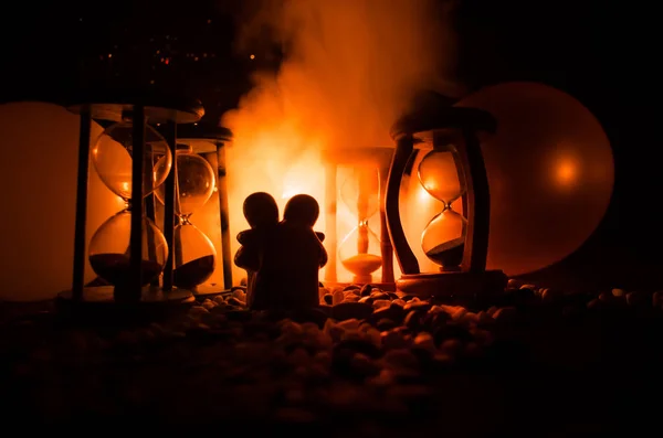 Tempo e conceito de amor. Silhuetas de figuras de cerâmica de brinquedo abraçando entre ampulhetas em fundo iluminado escuro com nevoeiro — Fotografia de Stock