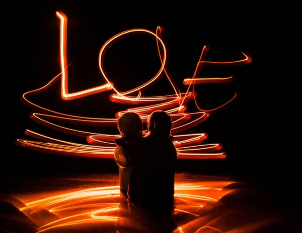 Two young lovers paint a heart on fire. Silhouette of couple and Love words on a dark background — Stock Photo, Image