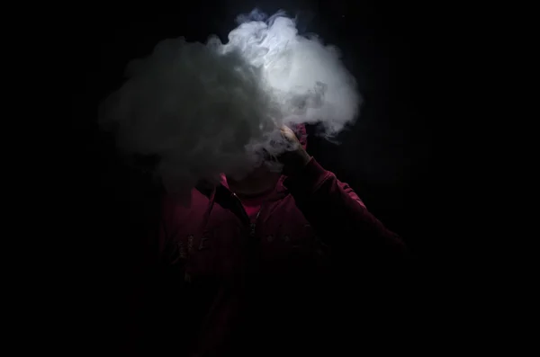 Hombre aspirante sosteniendo un mod. Una nube de vapor. Fondo negro. Vapeando un cigarrillo electrónico con mucho humo . — Foto de Stock