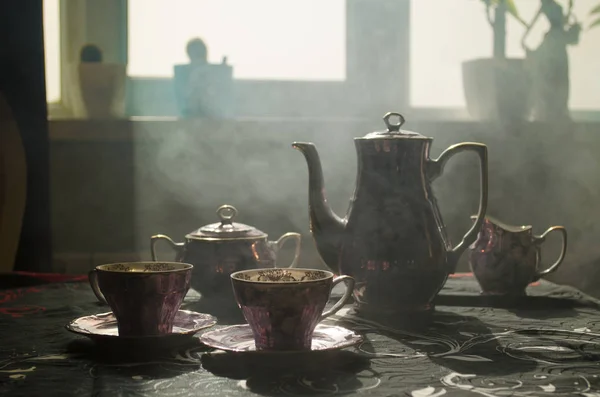 Пищевой фон чая и кофе тема. Старый старинный керамический чай или кофейник с чашками кувшин и сахарная чашка на темном фоне с огнями и дымом. Силуэты керамического чайника и чашки — стоковое фото