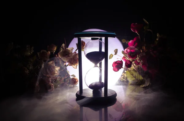 Concepto de tiempo. Arena que pasa a través de las bombillas de vidrio de un reloj de arena que mide el tiempo que pasa, ya que cuenta hasta un plazo. Silueta de clepsidras en humo sobre fondo oscuro. Con flores — Foto de Stock