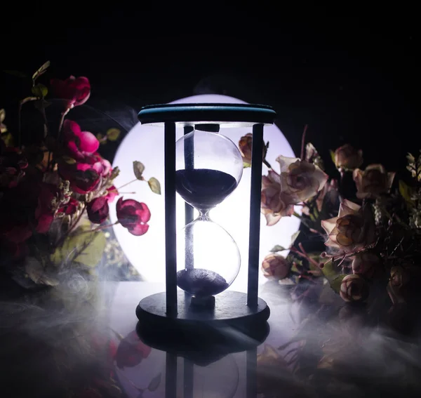 Tijd concept. Zand, passeren de glazen bollen van een zandloper de passerende tijd te meten, zoals het aftelt tot een datum. Silhouet van Hourglasses in rook op donkere achtergrond. Met bloemen — Stockfoto