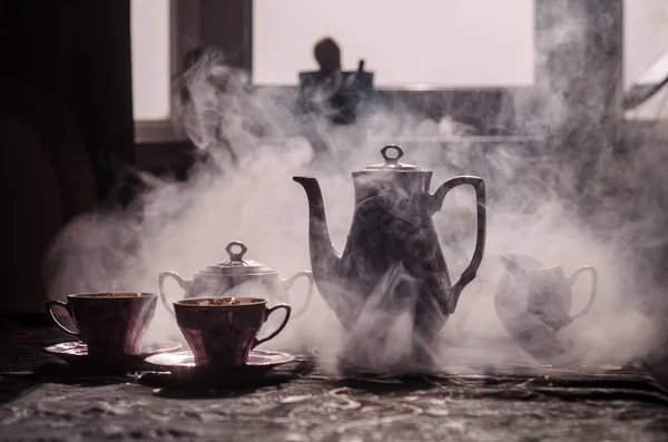 Comida fondo té y café tema. Tetera de cerámica antigua o cafetera con jarra de tazas y taza de azúcar sobre fondo oscuro con luces y humo. Siluetas de tetera y taza de cerámica — Foto de Stock