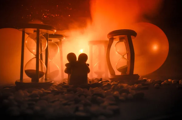 Concepto de tiempo y amor. Siluetas de figuras de cerámica de juguete abrazándose entre clepsidras en fondo oscuro iluminado con niebla — Foto de Stock