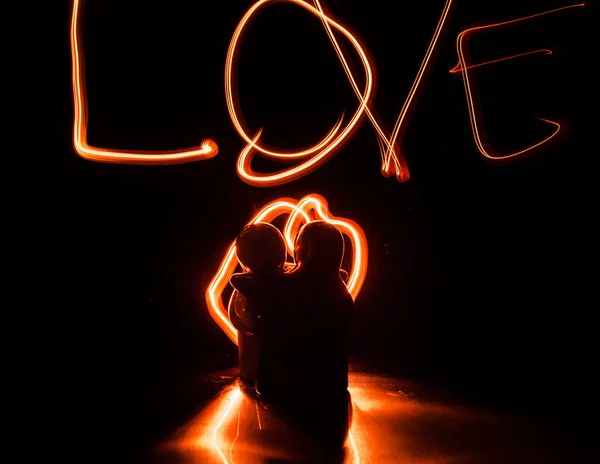 Two young lovers paint a heart on fire. Silhouette of couple and Love words on a dark background — Stock Photo, Image