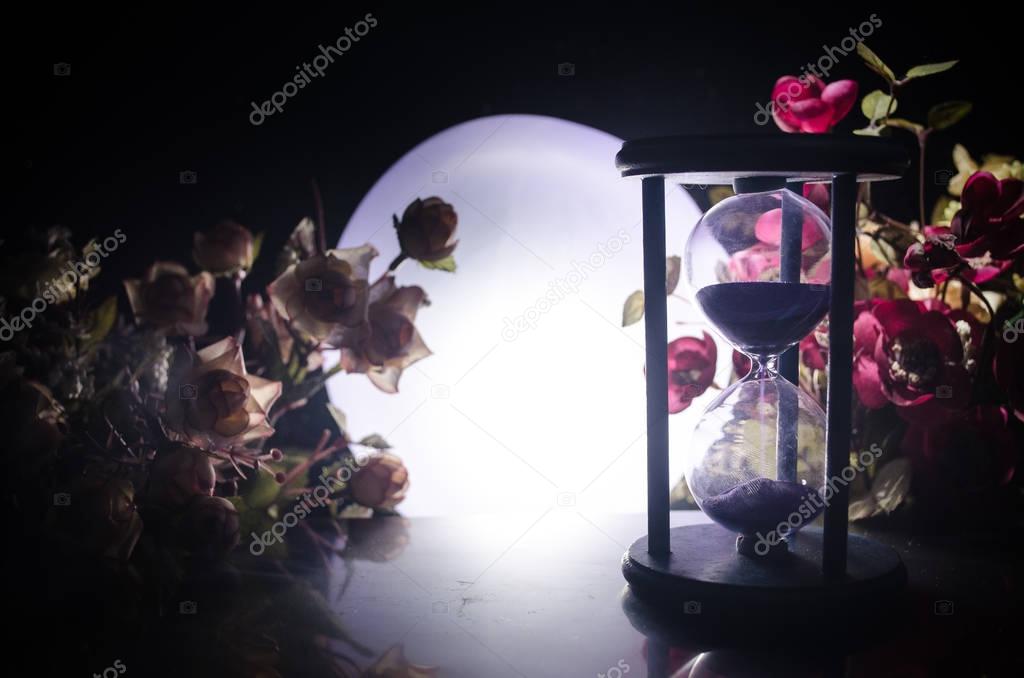 Time concept. Sand passing through the glass bulbs of an hourglass measuring the passing time as it counts down to a deadline. Silhouette of Hourglasses in smoke on dark background. With flowers