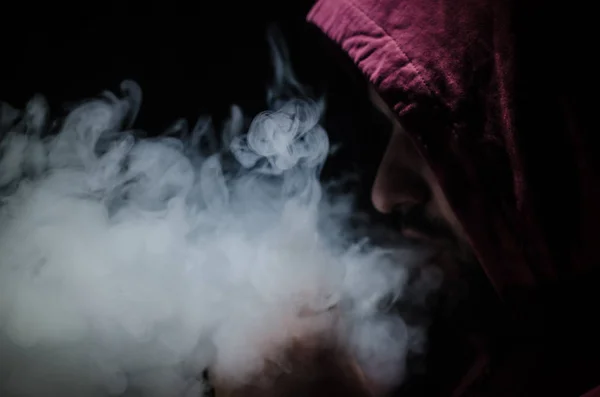 Hombre aspirante sosteniendo un mod. Una nube de vapor. Fondo negro. Vapeando un cigarrillo electrónico con mucho humo — Foto de Stock