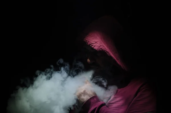 Hombre aspirante sosteniendo un mod. Una nube de vapor. Fondo negro. Vapeando un cigarrillo electrónico con mucho humo —  Fotos de Stock