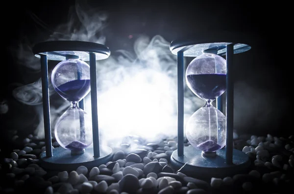 Time concept. Sand passing through the glass bulbs of an hourglass measuring the passing time as it counts down to a deadline. Silhouette of Hourglasses in smoke on dark background. With flowers — Stock Photo, Image