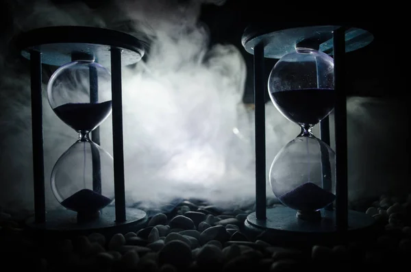 Time concept. Sand passing through the glass bulbs of an hourglass measuring the passing time as it counts down to a deadline. Silhouette of Hourglasses in smoke on dark background. With flowers — Stock Photo, Image