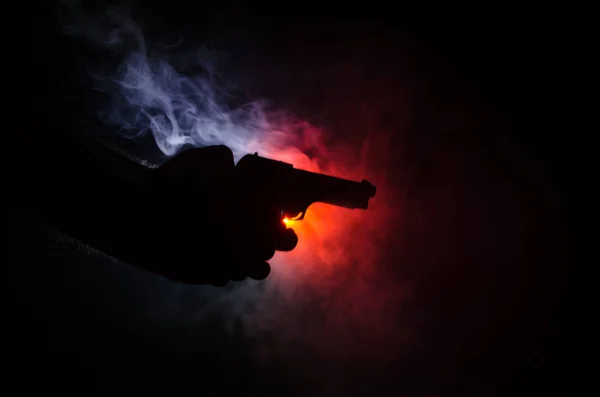 Mannenhand houden pistool op zwarte achtergrond met rook (geel oranje rood wit) terug gekleurde lichten, maffia moordenaar concept — Stockfoto