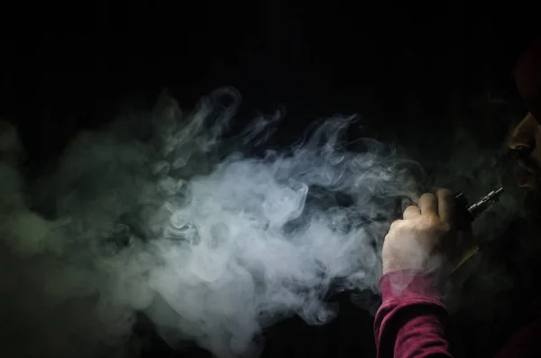 Homem a bater com um mod. Uma nuvem de vapor. Fundo preto. Vaping um cigarro eletrônico com muita fumaça — Fotografia de Stock