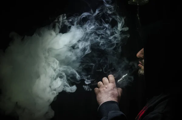 Hombre aspirante sosteniendo un mod. Una nube de vapor. Fondo negro. Vapeando un cigarrillo electrónico con mucho humo —  Fotos de Stock