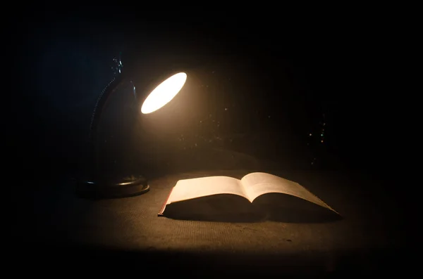 Buch neben glühender Tischlampe auf dunklem Hintergrund aufschlagen, Lampe und aufgeschlagenes Buch mit Rauch auf Hintergrund. surreal — Stockfoto