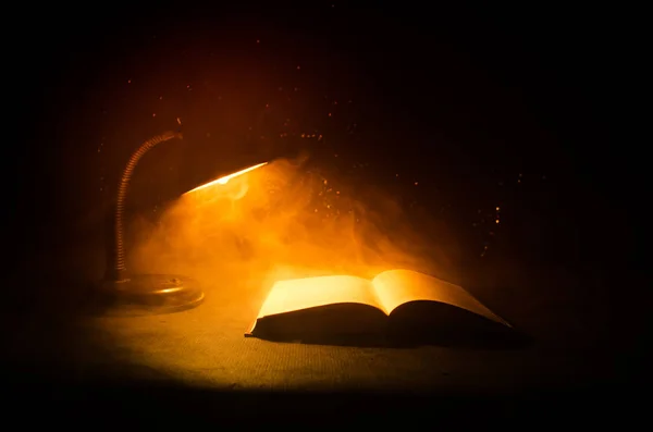 Ανοίξτε το βιβλίο κοντά σε πυρακτωμένο λαμπτήρα πίνακα σε σκούρο φόντο, λάμπα και άνοιξε βιβλίο με καπνό σε φόντο. Σουρεαλιστικά — Φωτογραφία Αρχείου
