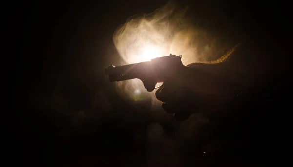 Mannenhand houden pistool op zwarte achtergrond met rook (geel oranje rood wit) terug gekleurde lichten, maffia moordenaar concept — Stockfoto