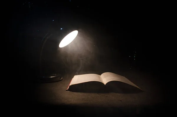 Buch neben glühender Tischlampe auf dunklem Hintergrund aufschlagen, Lampe und aufgeschlagenes Buch mit Rauch auf Hintergrund. surreal — Stockfoto