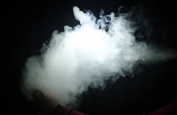 Hombre aspirante sosteniendo un mod. Una nube de vapor. Fondo negro. Vapeando un cigarrillo electrónico con mucho humo — Foto de Stock