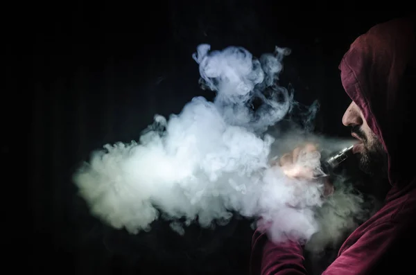 Homem a bater com um mod. Uma nuvem de vapor. Fundo preto. Vaping um cigarro eletrônico com muita fumaça — Fotografia de Stock