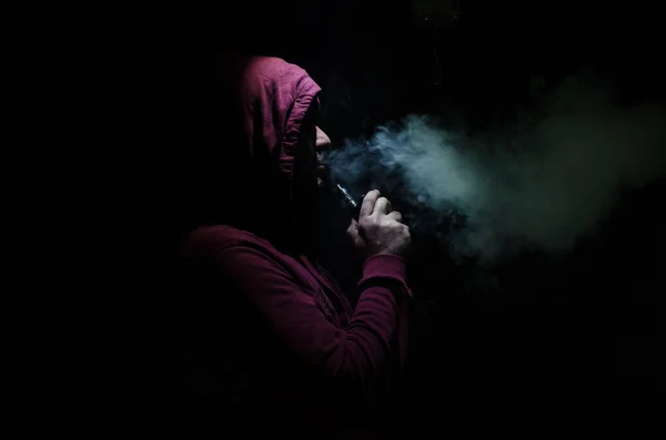 Hombre aspirante sosteniendo un mod. Una nube de vapor. Fondo negro. Vapeando un cigarrillo electrónico con mucho humo — Foto de Stock