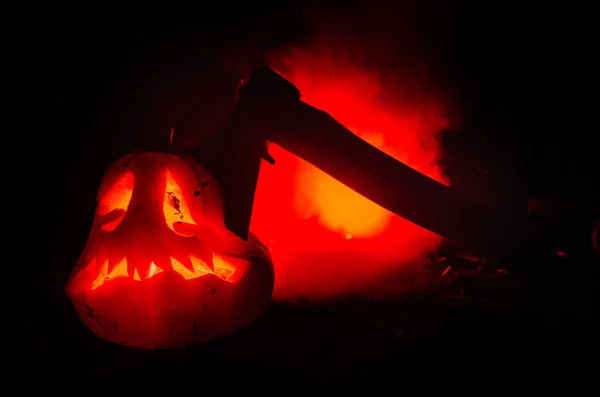 Scary orange pumpkin with carved eyes and a smile with burning candles and an ax on a dark background with fire sky. For the Halloween party. Empty space. — Stock Photo, Image