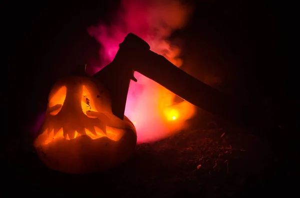 Zucca arancione spaventosa con occhi scolpiti e un sorriso con candele accese e un'ascia su uno sfondo scuro con cielo di fuoco. Per la festa di Halloween. Spazio vuoto . — Foto Stock