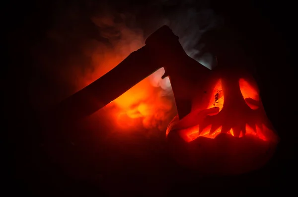 Abóbora laranja assustadora com olhos esculpidos e um sorriso com velas em chamas e um machado em um fundo escuro com céu de fogo. Para a festa de Halloween. Espaço vazio. . — Fotografia de Stock