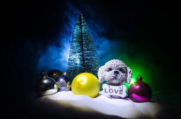 Toy dog - a symbol of the new year under the snow against the background of fir branches. Toy\'s dog as a symbol of 2018 New Year with a Christmas attributes the new year\'s inscription.