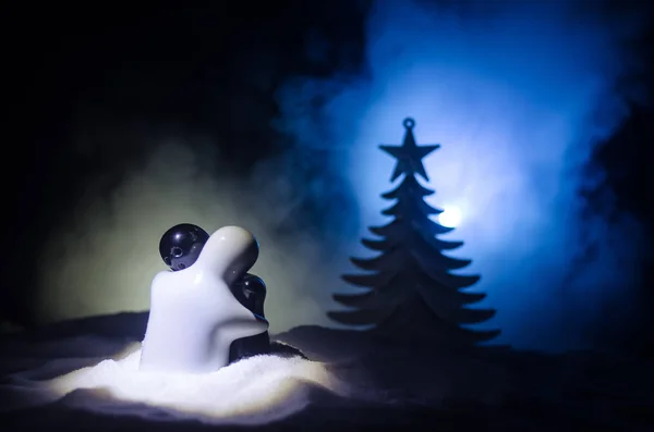 Love New Year Konzept. Mädchen und Junge aus Keramik umarmen sich, stehen auf dem weißen Schnee und farbigen Herzen und Weihnachtsbäumen auf verschwommenem, dunklem Hintergrund. Leere Räume. — Stockfoto