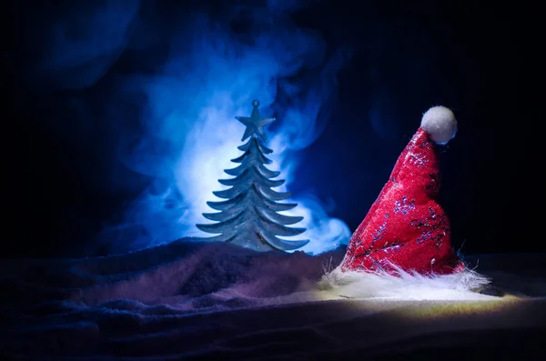 Christmas holiday New Year background with Santa Clause hat and blurred Christmas tree on snowy background. New Year conceptual image decoration with holiday attributes. Dark Toned background