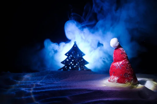 Christmas holiday New Year background with Santa Clause hat and blurred Christmas tree on snowy background. New Year conceptual image decoration with holiday attributes. Dark Toned background