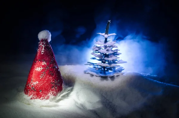 Christmas holiday New Year background with Santa Clause hat and blurred Christmas tree on snowy background. New Year conceptual image decoration with holiday attributes. Dark Toned background