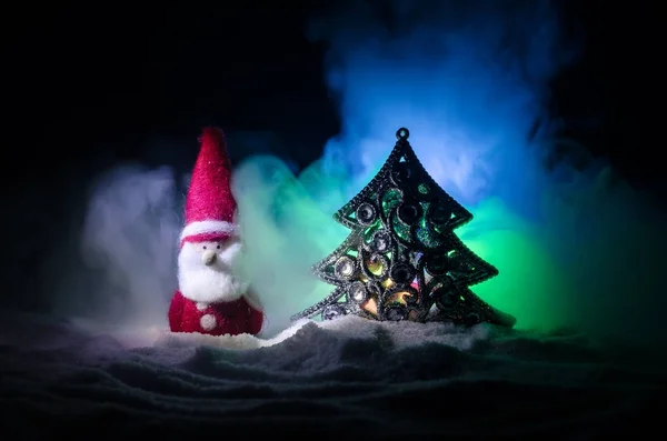 Feliz muñeco de Papá Noel en Navidad con árbol y nieve. Fondo bokeh colorido. Santa Claus y Feliz Navidad modelo figura juguete en oscuro tonificado niebla fondo — Foto de Stock