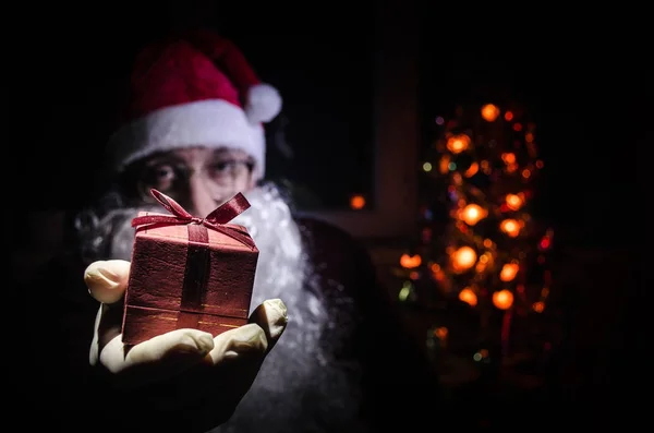 Santa Claus holding gift with festive holiday background. Santa Claus holding and offering a gift on his hand. Selective focus. Dark toned background — Stock Photo, Image