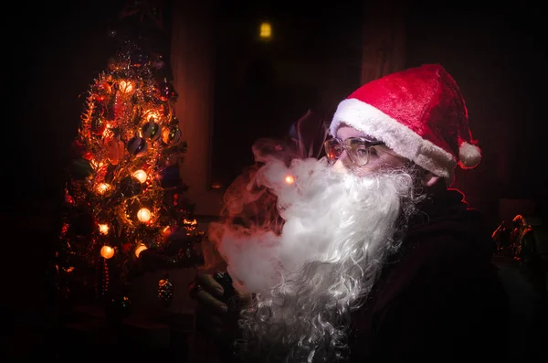 Śnięty Mikołaj vaping elektroniczny papieros ubrany jak tradycyjne Santa na ciemnym tle stonowanych z vape chmury. Selektywny fokus — Zdjęcie stockowe