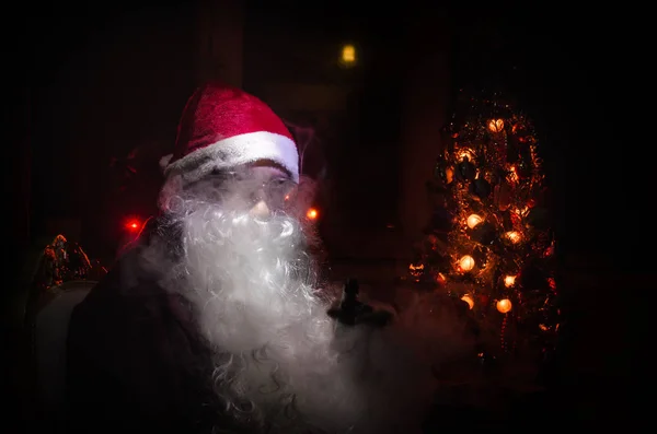 Śnięty Mikołaj vaping elektroniczny papieros ubrany jak tradycyjne Santa na ciemnym tle stonowanych z vape chmury. Selektywny fokus — Zdjęcie stockowe