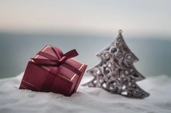Caja de regalo roja de Navidad y abeto en la nieve. Decoración de la casa de Navidad con nieve y árbol sobre fondo borroso durante el día con espacio para copiar. Enfoque selectivo . — Foto de Stock