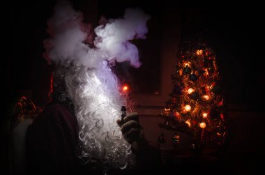 Noel Baba vaping elektronik sigara vape bulutlar karanlık bir tonda zemin üzerine geleneksel Noel Baba gibi giyinmiş. Seçici odak. Arka köknar ağacı