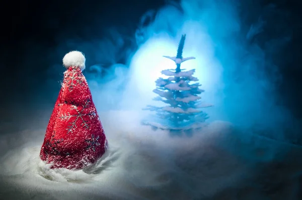 Christmas holiday New Year background with Santa Clause hat and blurred Christmas tree on snowy background. New Year conceptual image decoration with holiday attributes. Dark Toned background with fog