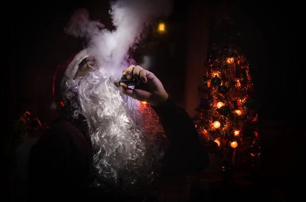 Santa Clause vaping electronic cigarette dressed as traditional Santa on a dark toned background with vape clouds. Selective focus