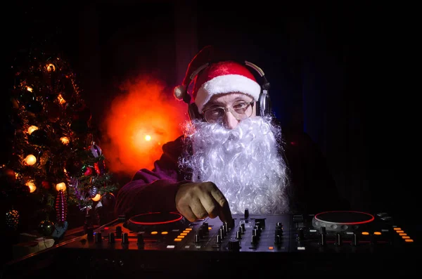 DJ Santa Claus mixing up some Christmas cheer. Dark disco club toned background. New Year's Eve event in the rays of light. Useful as poster. Selective focus — Stock Photo, Image