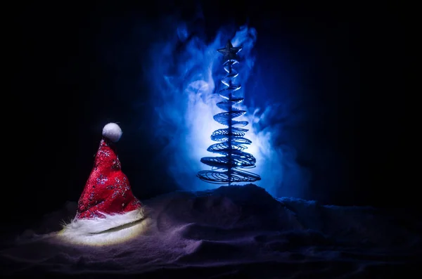 Christmas holiday New Year background with Santa Clause hat and blurred Christmas tree on snowy background. New Year conceptual image decoration with holiday attributes. Dark Toned background