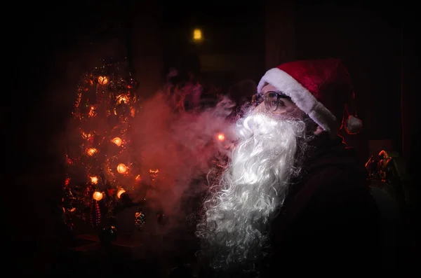 Śnięty Mikołaj vaping elektroniczny papieros ubrany jak tradycyjne Santa na ciemnym tle stonowanych z vape chmury. Selektywny fokus — Zdjęcie stockowe