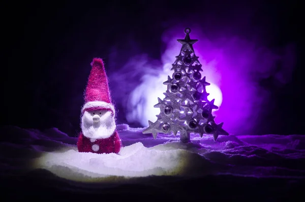 圣诞快乐圣诞老人娃娃在圣诞树和雪的时间。丰富多彩的散背景。圣诞老人和快乐的圣诞模型图玩具在黑暗的色调雾背景 — 图库照片